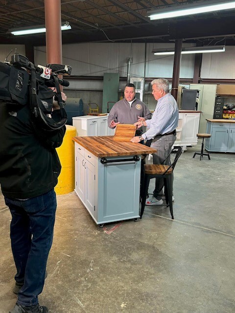 Wood You Build It founder Justin Robichaud explaining features of a handcrafted kitchen island to news anchor Doug Meehan while being filmed by a cameraman in a workshop.